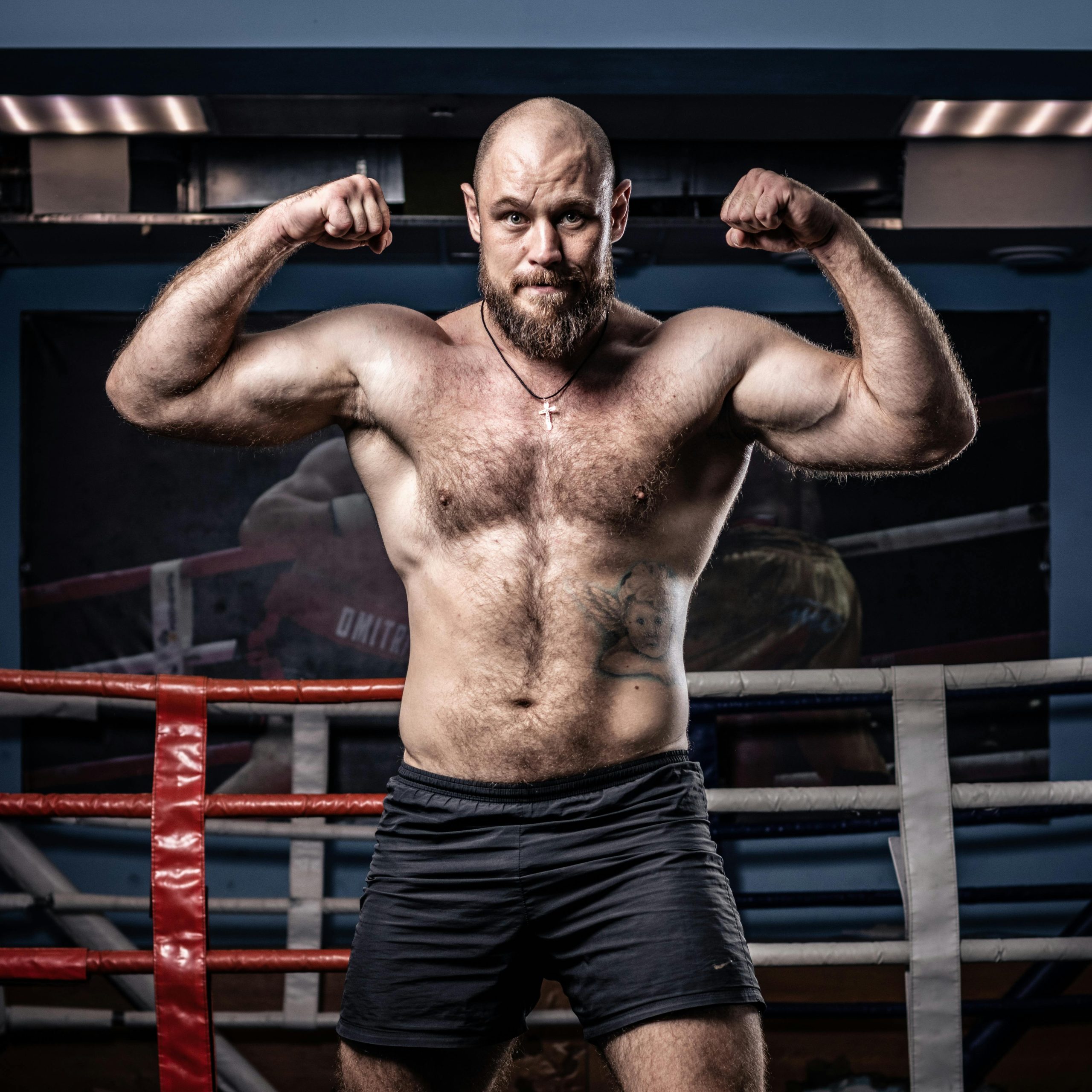 Strong muscular boxer posing with arms flexed in a gym ring, showcasing strength and fitness.